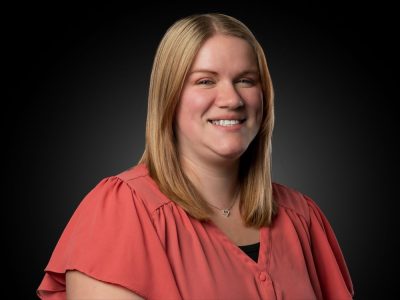 Smiling woman with shoulder-length blond hair in a coral blouse, posing against a dark studio background.
