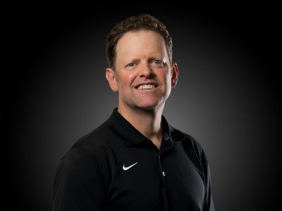Professional headshot of a smiling man in a black polo shirt against a dark gradient background, studio lighting.
