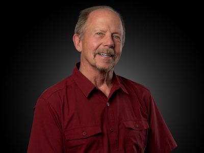 Smiling older man with gray hair in a maroon button-down shirt against a dark studio backdrop