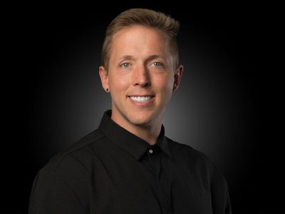 Head-and-shoulders portrait of a smiling man in a black shirt against a dark studio background, suitable for a corporate profile.
