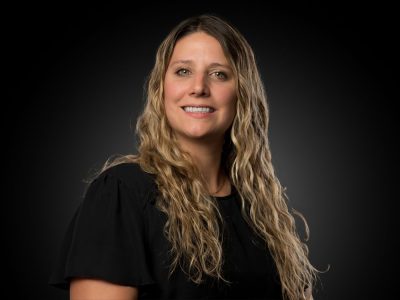 Portrait of a smiling woman with long wavy blonde hair wearing a black blouse against a dark gradient background in a studio portrait.