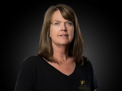Professional headshot of a middle-aged woman with shoulder-length brown hair, wearing a black V-neck shirt with a Village Properties logo visible on the chest.