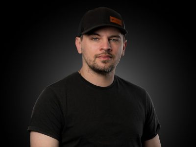 Portrait of a man in a black t-shirt and black cap against a dark studio backdrop with a subtle vignette.