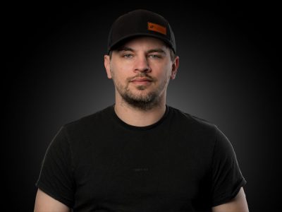 Head-and-shoulders portrait of a man in a black T-shirt and black cap against a dark gradient background.