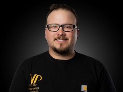 Portrait of a man with glasses and a goatee, wearing a black shirt with a VP Village Properties logo, smiling against a dark gradient background