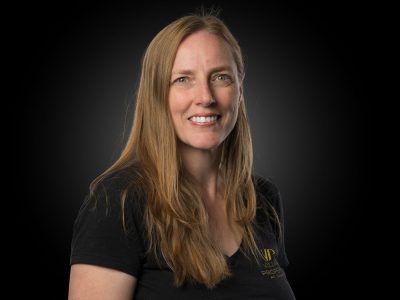 Portrait of a smiling woman with long blonde hair, wearing a black t-shirt, against a dark studio background.