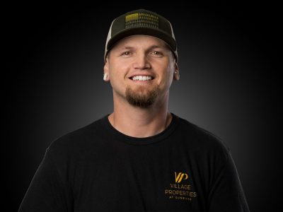 Portrait of a smiling man in a black Village Properties t-shirt and cap, against a dark gradient background.