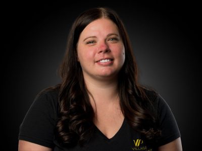 Portrait of a smiling woman with long dark hair, wearing a black V-neck shirt with a small yellow logo, against a dark studio background