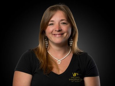 Portrait of a smiling woman with shoulder-length light brown hair, wearing a black V-neck shirt with a Village Properties logo on the chest and a white necklace.