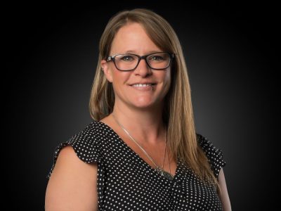 Portrait of a smiling woman with glasses wearing a black polka dot blouse, against a dark studio background.