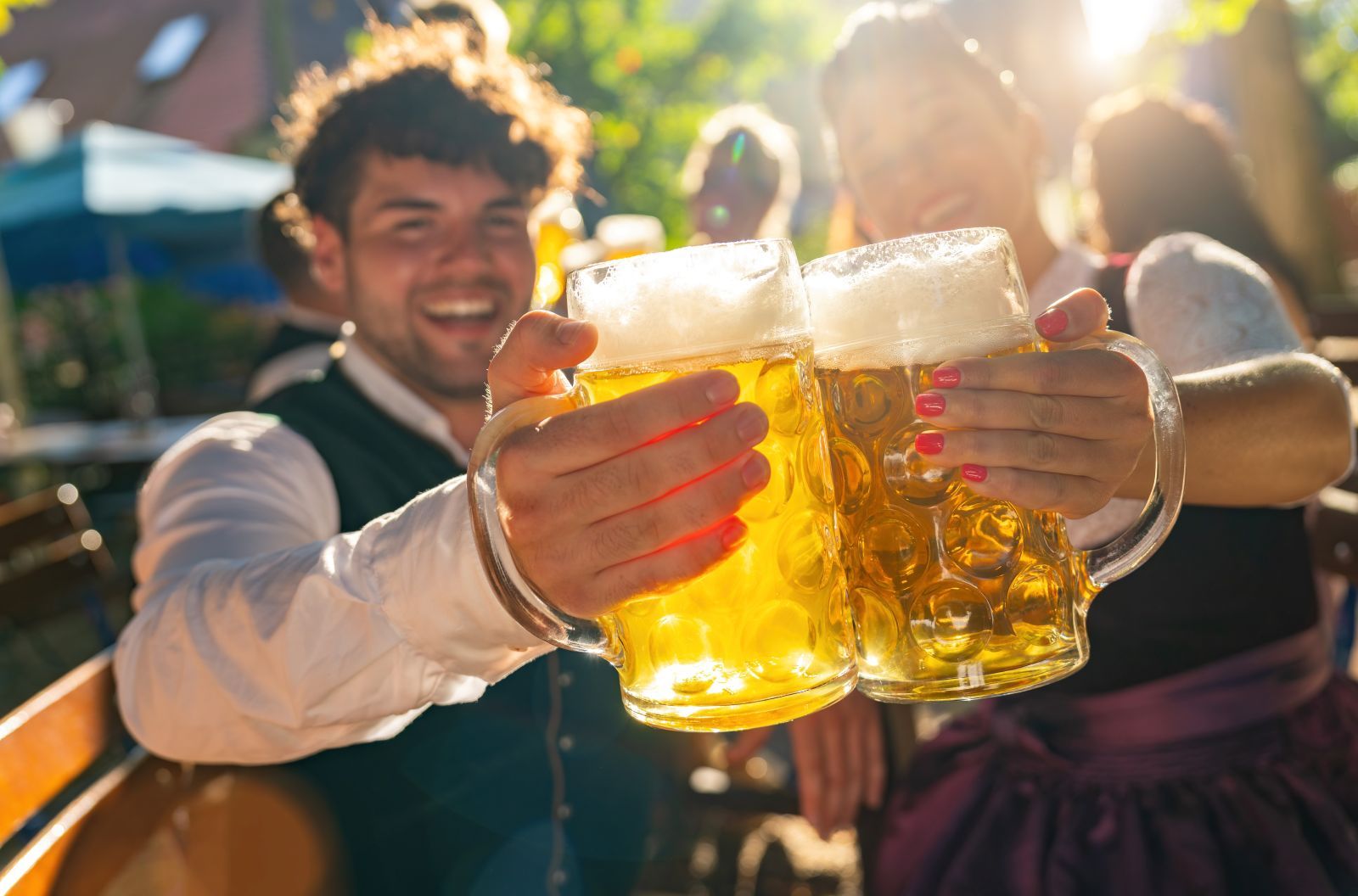 Friends toasting with large beer mugs outdoors on a sunny day, smiles and clinking glasses.
