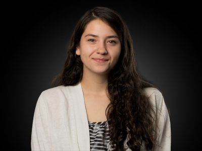 Portrait of a young woman with long dark hair, smiling, wearing a cream cardigan over a striped top, against a dark studio background.