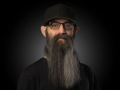 Portrait of a man with a long gray beard, glasses, and a black cap, wearing a black shirt against a dark studio background.