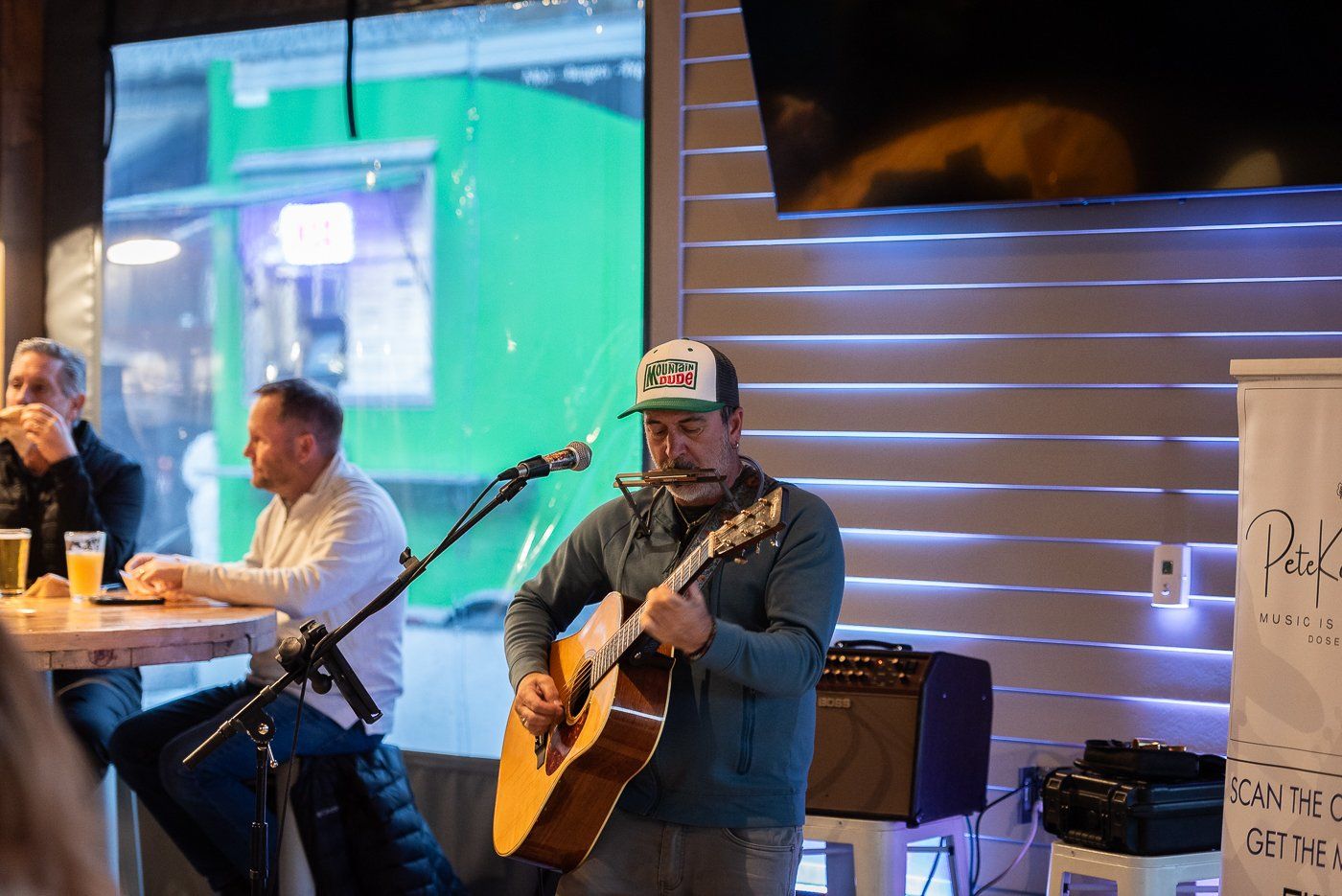 Man wearing a Mountain Dew-style hat plays an acoustic guitar and sings into a mic at a small indoor venue.